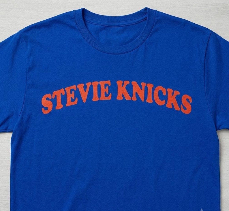 May include: A bright blue short-sleeved t-shirt featuring the text STEVIE KNICKS printed in a bold, arched orange font across the chest. The garment has a classic crew neckline and is made of a solid-colored fabric with a smooth texture.