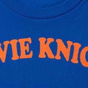 May include: A royal blue short-sleeved t-shirt featuring the text STEVIE KNICKS printed in a bold, arched orange font across the chest. The garment has a standard crew neckline and a smooth, solid-colored fabric construction throughout the front.
