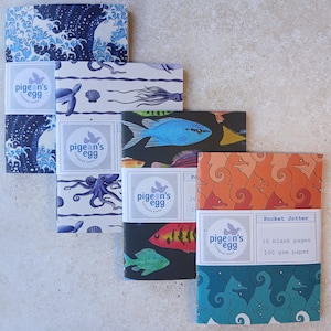 May include: Four rectangular pocket jotter notebooks with paper bands featuring the pigeon's egg logo. Designs include blue ocean waves, marine life with octopuses, colourful tropical fish on black, and orange and teal seahorse patterns.