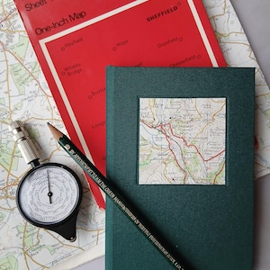 May include: A dark green hardback notebook featuring a square map inlay on the cover, resting on a red Buxton & Matlock map. A black pencil with gold lettering and a circular silver and black map measuring tool are positioned on the map surface.