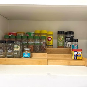 Peut inclure: Étagère de placard de cuisine remplie de divers pots et contenants d'épices. Les épices sont disposées sur deux présentoirs en bambou à plusieurs niveaux. Les marques incluent Old Bay, Tex Joy et HEB. Les pots sont remplis d'herbes et d'épices.