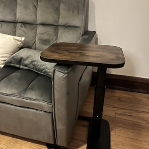 May include: A dark brown, square-topped side table with a black, adjustable-height base. The table is positioned next to a gray, tufted sofa. The table's surface has a wood grain appearance, and the base is a simple, modern design.