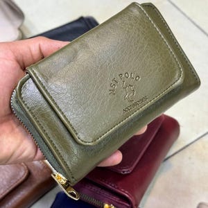 Op de afbeelding: Olijfgroene leren portemonnee met ritssluiting en flap. De portemonnee heeft de in reliëf aangebrachte tekst "MST POLO ACCESSORIES" en een goudkleurige ritssluiting. Andere portemonnees in verschillende kleuren zijn zichtbaar op de achtergrond.