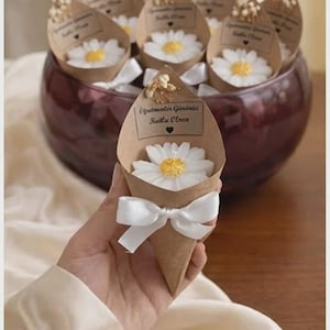 Könnte beinhalten: Eine Sammlung kegelförmiger brauner Papierverpackungen, die jeweils eine weiße gänseblümchenförmige Kerze mit gelber Mitte enthalten. Jedes Päckchen ist mit einem weißen Band gebunden, mit Trockenblumen verziert und trägt ein Etikett mit dem Text "Öğretmenler Gününüz Kutlu Olsun".