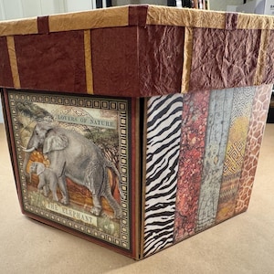 May include: A decorative box with a brown, textured lid and sides featuring various patterns. One side displays an elephant illustration with the text "LOVERS OF NATURE" and "THE ELEPHANT". The box has a rectangular shape.