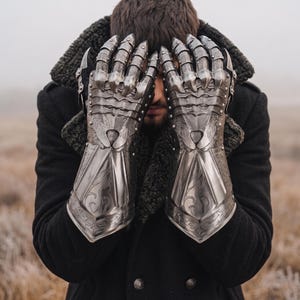 May include: A pair of silver-colored, articulated metal gauntlets with intricate engraved designs. The gauntlets feature pointed finger extensions and are worn over a black coat with a fur collar. The background is a blurred outdoor scene.