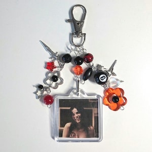 May include: A keyring featuring a square clear plastic frame containing a photograph of a person with dark hair, surrounded by dangling charms including a silver dagger, a black eight ball, red and orange flower beads, and star shapes.