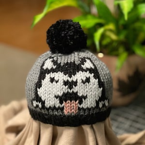 May include: A gray and black knitted hat with a black pom-pom on top. The hat features a white and black knit design that resembles a face with a pink nose.