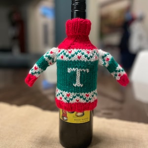 May include: A green, white, and red knitted bottle cozy with a red pom pom on top. The cozy has a white letter "T" on the front and a red heart pattern. The cozy is on a wine bottle with a label that says "Red Wine".