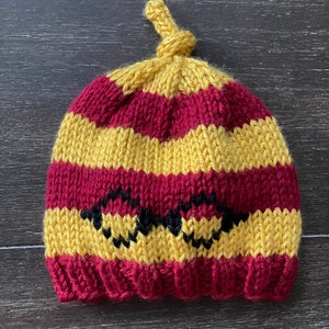 Peut inclure: Un bonnet tricoté avec des rayures jaunes et rouges et un motif en zigzag noir. Le bonnet a un nœud en haut.