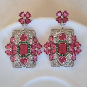 Può includere: Un paio di orecchini pendenti color argento con una pietra rossa rettangolare centrale circondata da piccole pietre verdi. Il design include diverse pietre rosa a forma di cuore e cristalli trasparenti su una cornice rettangolare geometrica.