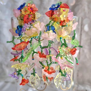 Op de afbeelding: Een paar statement oorbellen met een dichte tros van veelkleurige, doorschijnende acrylbloemen en groene glaskralenranken. Het bloemstuk bevat tinten roze, geel, rood, blauw en paars, wat zorgt voor een levendig, gelaagd botanisch ontwerp met bungelende bladvormige accenten.