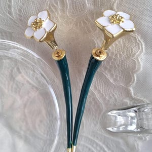 Puede incluir: Dos horquillas para el pelo con flores blancas y centros dorados. Las flores tienen bordes dorados. Los tallos son de color verde azulado oscuro y terminan en puntas doradas. Accesorios para el cabello sobre un fondo texturizado.