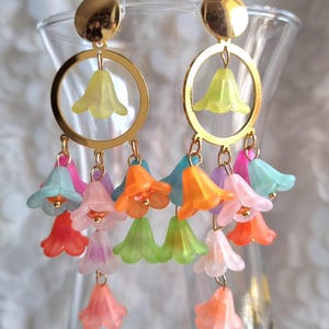 Peut inclure: Boucles d'oreilles dorées avec un motif circulaire et une fleur jaune au centre. En dessous, plusieurs brins de breloques de fleurs translucides colorées dans les tons rose, orange, vert et bleu pendent des boucles d'oreilles. Les boucles d'oreilles sont sur un fond flou.