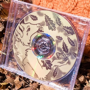 Op de afbeelding: Een doorzichtig plastic CD-doosje met een schijfje waarop een illustratie staat van een vosachtig wezen dat een vlinder vasthoudt, omringd door vallende eikenbladeren. Het kunstwerk is uitgevoerd in tinten beige en donkerbruin.