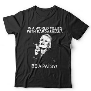 May include: A black short-sleeved t-shirt featuring a white graphic print of a person smoking a cigarette. The text above the graphic reads IN A WORLD FILLED WITH KARDASHIANS and the text below the graphic reads BE A PATSY! in white capital letters.