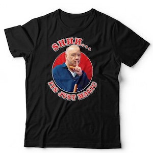 May include: A black t-shirt featuring a circular graphic of a person in a blue shirt holding a finger to their lips in a shushing gesture. The text SHHH... IT'S JUST MAGIC is printed in red with a white outline around the circular image.