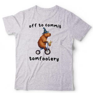 May include: A light grey t-shirt featuring a graphic of a brown capybara wearing a blue wizard hat with yellow stars, riding a blue tricycle with a small basket, accompanied by the black text "off to commit tomfoolery" above and below the graphic.