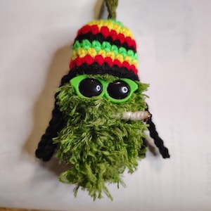 Pode incluir: Um boneco de crochê verde e felpudo com um gorro de listras horizontais pretas, vermelhas, amarelas e verdes. O boneco usa óculos de sol de plástico verde brilhante e um pequeno objeto branco enrolado sai de sua boca.