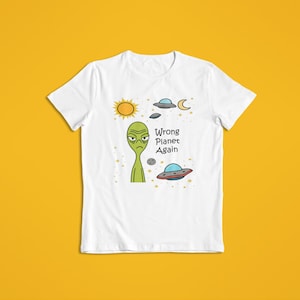 Puede incluir: Camiseta blanca con un diseño de un alienígena de dibujos animados con expresión de enfado, rodeado de un sol, una luna, estrellas y platillos volantes. El texto "Wrong Planet Again" está impreso debajo.