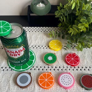 May include: A collection of circular silicone fruit-themed beverage can covers and a matching coaster. The set includes designs resembling lime, lemon, kiwi, grapefruit, coconut, orange, dragon fruit, and watermelon, displayed with a soda can.