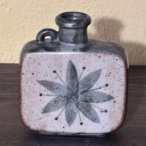 Pode incluir: Um vaso de cerâmica de formato quadrado com gargalo estreito e uma pequena alça lateral. O corpo apresenta um acabamento salpicado em branco sujo com um padrão central de estrela ou flor cinza. O gargalo e a alça têm um esmalte cinza escuro.