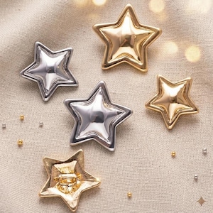 Op de afbeelding: Een assortiment stervormige decoraties in zilver- en goudtinten. De sterren zijn gerangschikt op een neutraal gekleurde stof, met een paar kleine, ronde versieringen verspreid. De metalen sterren hebben een glanzend, reflecterend oppervlak.