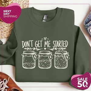 May include: An olive green crewneck sweatshirt featuring a white graphic print of three glass jars with varying levels of contents. The text above the jars reads "DON'T GET ME STARTED" in a stylized font, accented by small heart and leaf illustrations.
