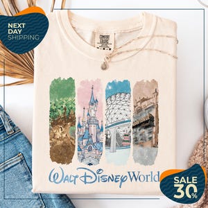A white t-shirt with a graphic design featuring illustrations of various Disney landmarks, including the castle, the Eiffel Tower, and the Hollywood sign. The t-shirt is displayed on a white background, and there are jeans and a sweater visible in the background.