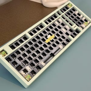 May include: A pale green mechanical keyboard chassis featuring a dark gray switch plate, exposed white stabilizers, and a few installed green switches. A small piece of yellow masking tape is applied to the center of the plate.