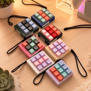 Pode incluir: Sete teclados macro quadrados com nove teclas mecânicas coloridas cada, com o nome da marca MiloMech impresso na lateral. Cada dispositivo inclui uma alça de pulso preta e exibe uma combinação de cores única de teclas em tons pastel e vibrantes.