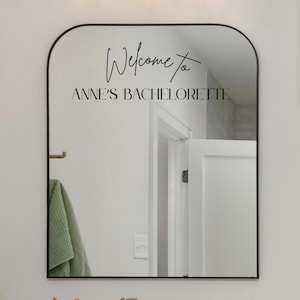 May include: An arched wall mirror with a thin black frame featuring black vinyl lettering on the glass surface. The text reads Welcome to ANNE'S BACHELORETTE in a combination of cursive script and bold, capitalized sans-serif typography.