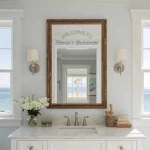 May include: A bathroom mirror with a wooden frame, reflecting a view of the sea. The mirror has the text "WELCOME TO Hannah's Bachelorette" in black script. The vanity has a marble countertop, a sink, and a vase of white flowers.