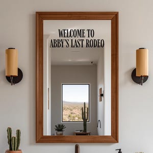 May include: A rectangular wall mirror with a medium-toned wooden frame. The glass surface features black lettering that reads "WELCOME TO ABBY'S LAST RODEO". The mirror reflects a room with a window showing a desert landscape and a cactus.