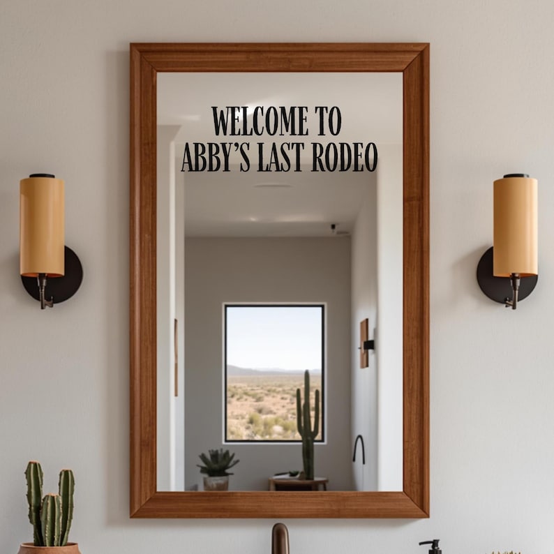 May include: A rectangular wall mirror with a medium-toned wooden frame. The glass surface features black lettering that reads "WELCOME TO ABBY'S LAST RODEO". The mirror reflects a room with a window showing a desert landscape and a cactus.