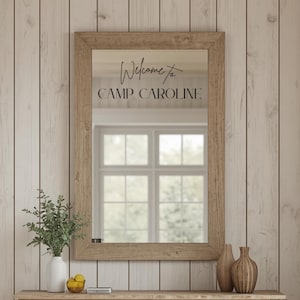 May include: A rectangular mirror with a light brown wood frame featuring the black text Welcome to CAMP CAROLINE printed on the glass surface. The mirror reflects a window and is positioned above a surface with a white vase and two wooden vases.