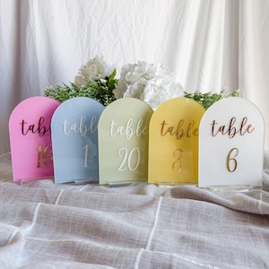Pode incluir: Cinco placas de números de mesa arqueadas em cores pastel: rosa, azul, verde, amarelo e branco. Cada placa tem a palavra "mesa" e um número em uma cor contrastante. As placas são exibidas em bases de acrílico transparente.