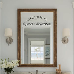 May include: A rectangular wall mirror with a dark brown wooden frame featuring black vinyl lettering on the glass that reads WELCOME TO Hannah's Bachelorette. The mirror reflects a bright room with a window looking out toward a beach scene.