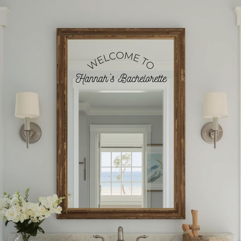 May include: A rectangular wall mirror with a dark brown wooden frame featuring black vinyl lettering on the glass that reads WELCOME TO Hannah's Bachelorette. The mirror reflects a bright room with a window looking out toward a beach scene.