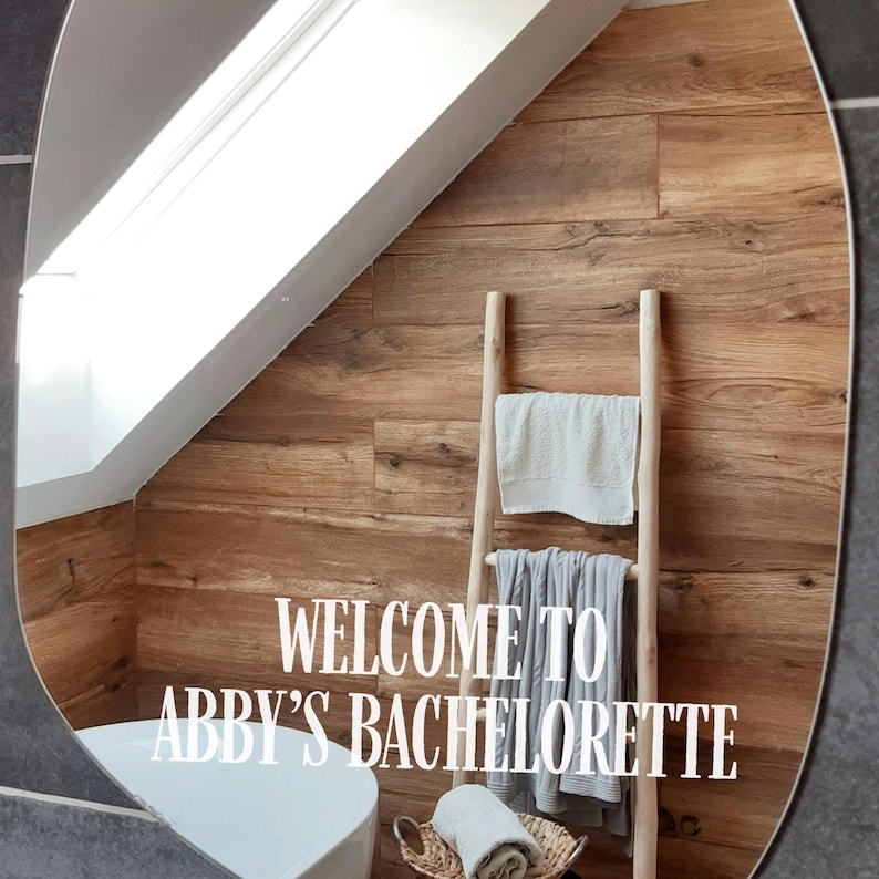 May include: An oval mirror featuring white vinyl lettering that reads "WELCOME TO ABBY'S BACHELORETTE". The mirror reflects a wooden wall, a light-colored wooden towel ladder with hanging towels, and a white bathtub in a bright, modern bathroom.