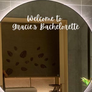 May include: A round mirror featuring white vinyl lettering that reads Welcome to Gracie's Bachelorette. The mirror has a thin black frame and is illuminated by a soft light, reflecting a dark wall with leaf-shaped wall decor and a bed.