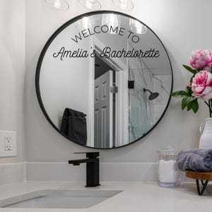 May include: A round mirror with a thin black frame featuring the black text WELCOME TO Amelia's Bachelorette applied to the glass surface. The mirror reflects a bathroom interior with a doorway, a shower head, and a dark towel hanging on a hook.