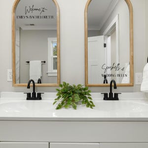 May include: Two arched mirrors with light wood frames mounted above a double vanity. The left mirror features black text reading Welcome to EMILY'S BACHELORETTE. The right mirror features black text reading Seashells & WEDDING BELLS.