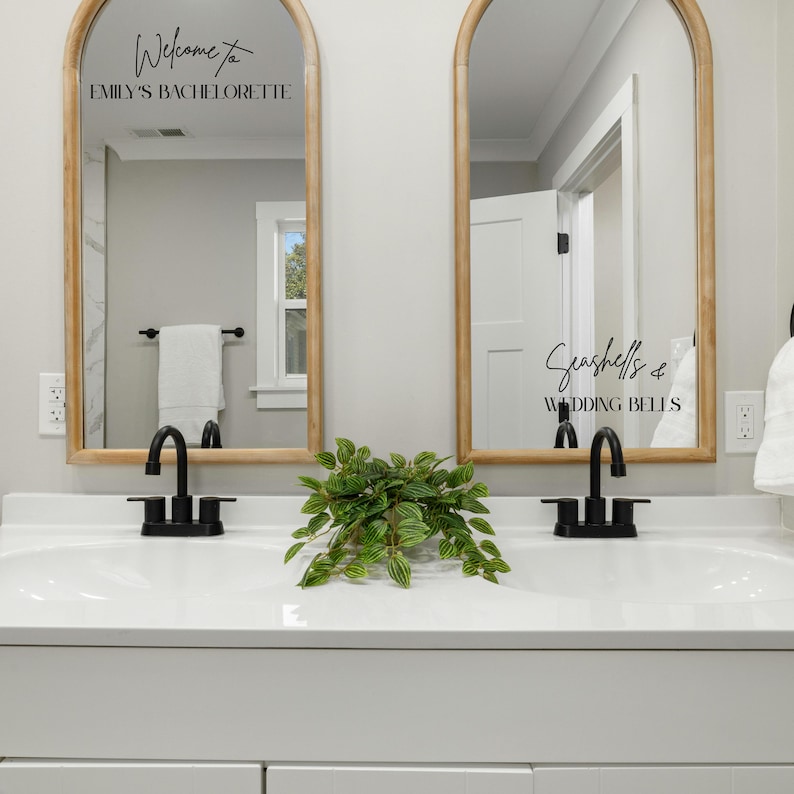 May include: Two arched mirrors with light wood frames mounted above a double vanity. The left mirror features black text reading Welcome to EMILY'S BACHELORETTE. The right mirror features black text reading Seashells & WEDDING BELLS.