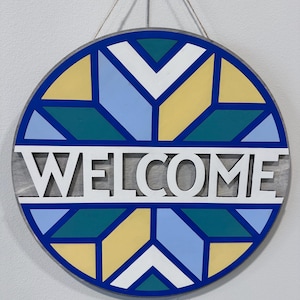 May include: A round wooden welcome sign with a geometric pattern in shades of blue, yellow, and green. The word "WELCOME" is cut out in white letters.