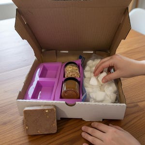 May include: An open cardboard box containing soap-making supplies. The box includes a purple mold, jars of ingredients, a bag of white cubes, and a bar of soap. A person's hand is reaching into the box.