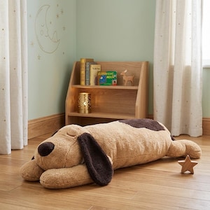 Op de afbeelding: Een groot, pluche hondvormig kussen in tinten bruin en beige, liggend op een houten vloer. De hond heeft flaporen en een donkerbruine vlek op zijn rug. Een houten ster en een boekenplank met boeken staan op de achtergrond.