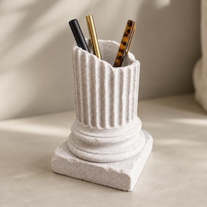 May include: A light grey, speckled stone pencil holder shaped like a broken classical column with a square base. The cylindrical top features vertical fluting and holds three pens: one matte black, one metallic gold, and one brown tortoiseshell pattern.