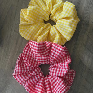 May include: Two oversized scrunchies. One is yellow and white gingham, the other is red and white gingham. The fabric is gathered around a hair tie. These accessories are perfect for adding a pop of color to any hairstyle.