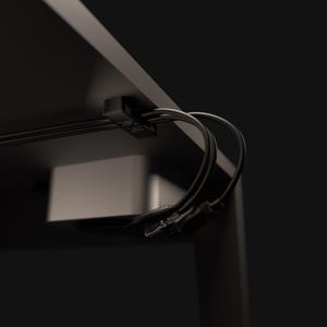 May include: A black cable tidy clip is fixed under a dark desk. Several black cables are neatly arranged and held by the clip. A silver device is visible in the background. The setup promotes a tidy workspace, with cables out of sight.